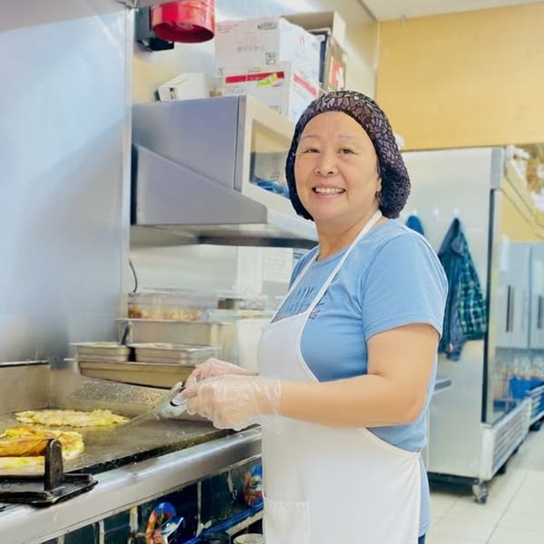 A smiling deli worker in a hairnet and apron cooks food on a griddle in a commercial kitchen.