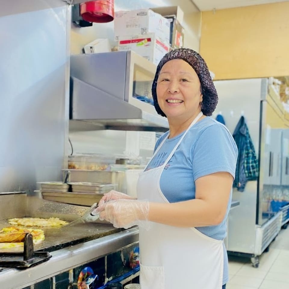 A smiling deli worker in a hairnet and apron cooks food on a griddle in a commercial kitchen.