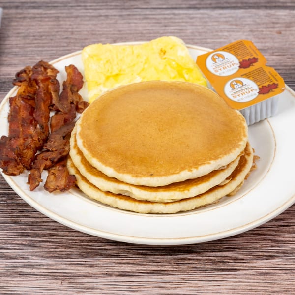 A breakfast plate with a stack of pancakes, scrambled eggs, crispy bacon, and two syrup packets.