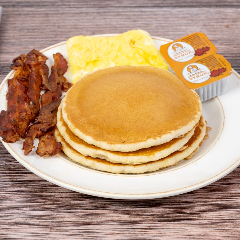 A breakfast plate with a stack of pancakes, scrambled eggs, crispy bacon, and two syrup packets.